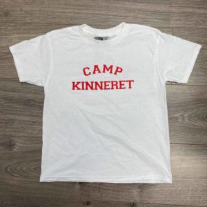 A white t-shirt with "Camp Kinneret" printed in red letters on the front, displayed on a wooden floor.