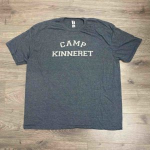 A grey t-shirt with the words "Camp Kinneret" printed in white lettering on the front, laid flat on a wooden floor.