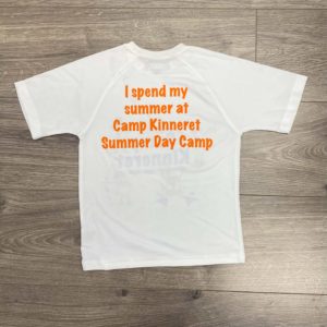 White t-shirt with orange text on the back reading, "I spend my summer at Camp Kinneret Summer Day Camp," lying flat on a wooden floor.