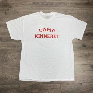 A white T-shirt with "Camp Kinneret" printed in red capital letters on the chest, laid flat on a wooden floor background.