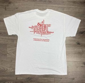 White t-shirt with a word cloud design on the back, featuring various event names. Text below reads: "Celebrating Over 60 Summers of Adventures and Memories!" The shirt is laid flat on a wooden surface.