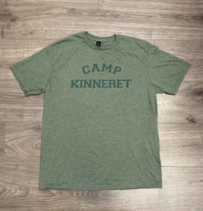 A green t-shirt laying flat on a wooden floor. The shirt has the words "CAMP KINNERET" printed in dark green text on the front.