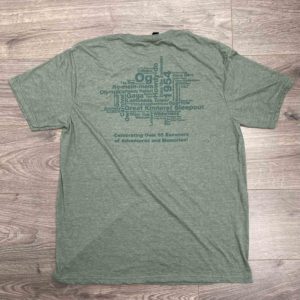 Back of a green T-shirt featuring a word cloud design with various adventure-related terms. Text below reads "Celebrating Over 85 Summers of Adventures and Memories!" on a wooden floor background.