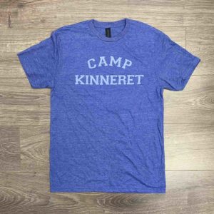 A blue T-shirt with the words "CAMP KINNERET" printed in white, laid flat on a wooden floor.