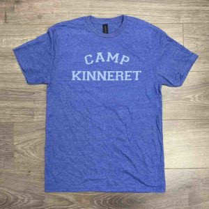 A blue t-shirt with the words "Camp Kinneret" printed in white letters on the front, laid flat on a wooden floor.