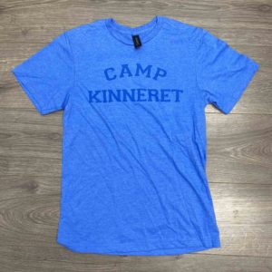 A blue T-shirt with the words "CAMP KINNERET" printed in dark blue letters lies flat on a wooden surface.