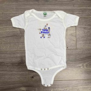 A white baby onesie with short sleeves and a graphic design in the center depicting animals with the text "Camp Kinneret" printed on it, laid flat on a wooden surface.