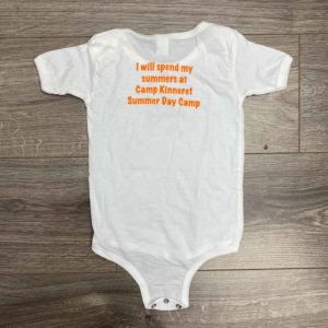 A white baby onesie with orange text stating, "I will spend my summers at Camp Kinneret Summer Day Camp," is laid flat on a wooden floor.