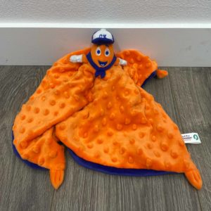 An orange plush blanket with a textured pattern, featuring a small toy mascot with a hat and blue outfit at the corner, is spread on a wooden floor against a white wall.