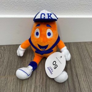 Orange plush toy with a blue and white cap labeled "C.K." sits on a wooden floor against a white wall. It has white gloves, shoes, and a bow tie, and holds a tag that reads "og I am an explorer".