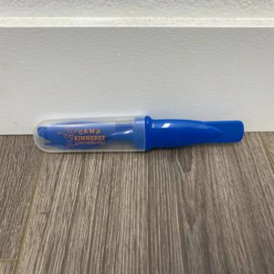 A plastic whistle in a clear protective case with a blue cap and a logo that reads "Camp Kinneret." The whistle is resting on a wooden floor against a white wall.