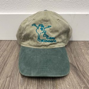 A beige and green baseball cap with an embroidered blue logo and text reading "Camp Kinneret" on it, placed on a wood floor against a white wall.